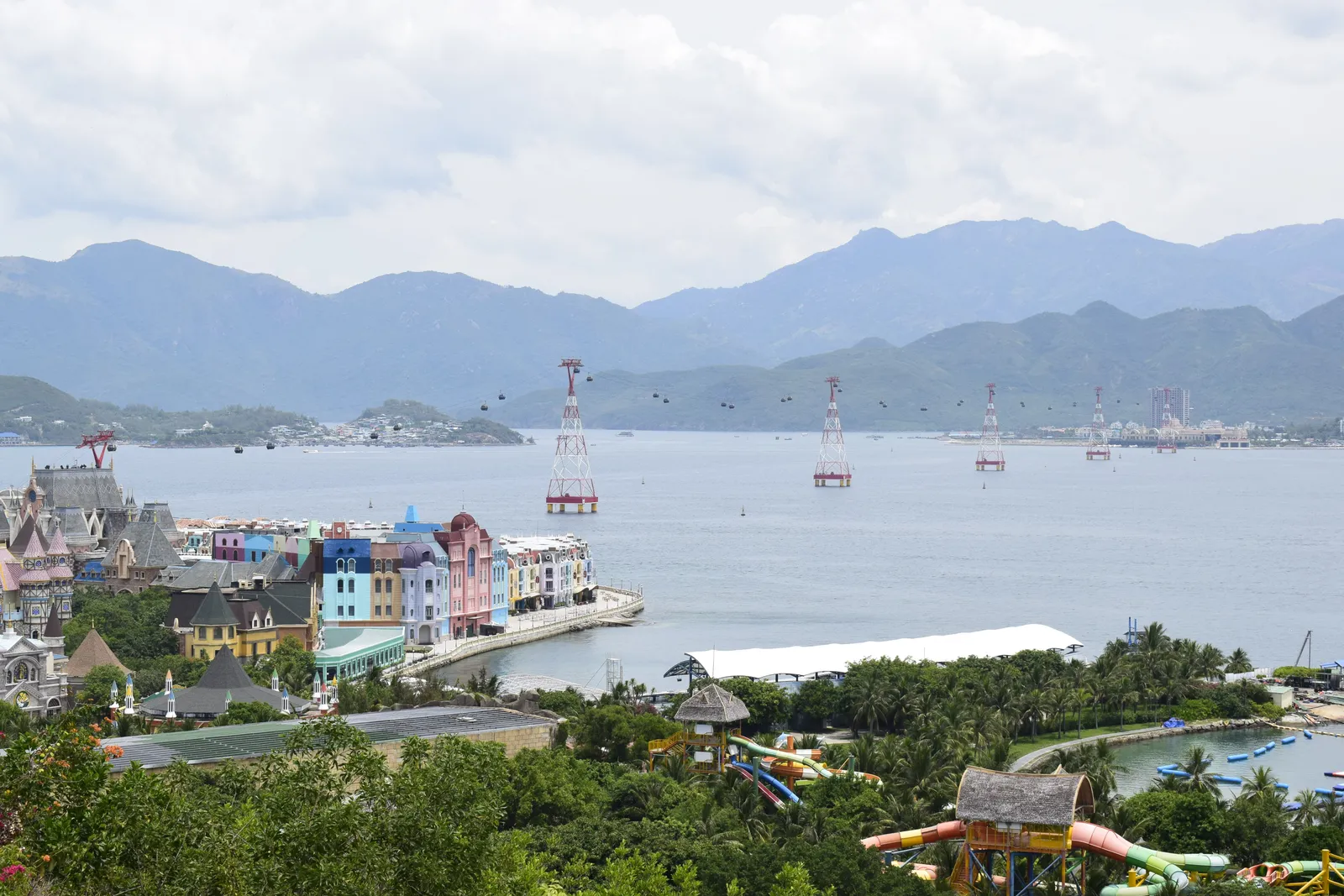 Nha Trang Bay seen from a hillside, with a brightly coloured fairytale-style waterfront, cable-car towers crossing the water and mountains behind