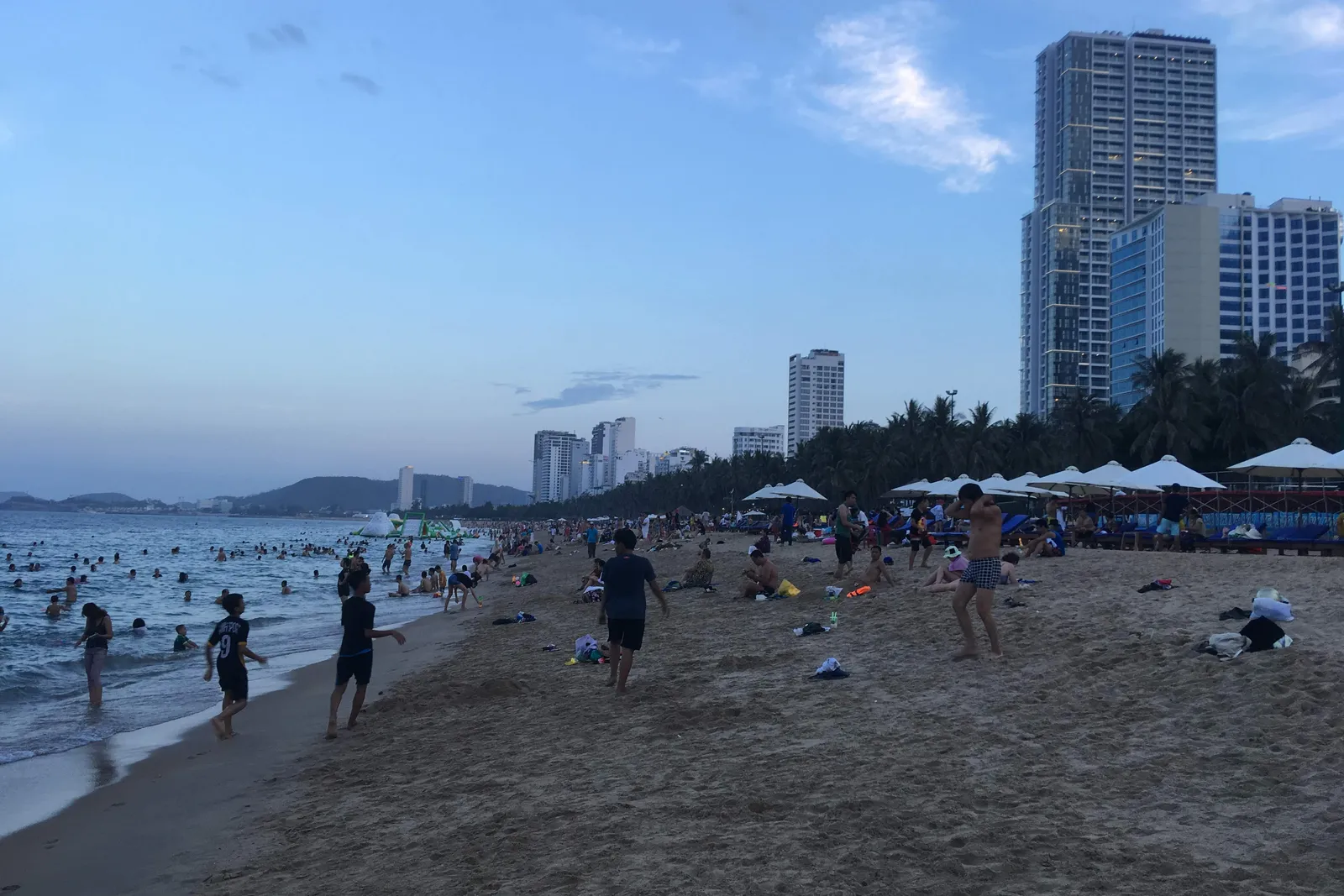 Ludzie spacerujący i wiosłujący na szerokiej piaszczystej plaży o zmierzchu, z parasolami plażowymi, palmami i wysokimi wieżami hotelowymi w tle, w Nha Trang
